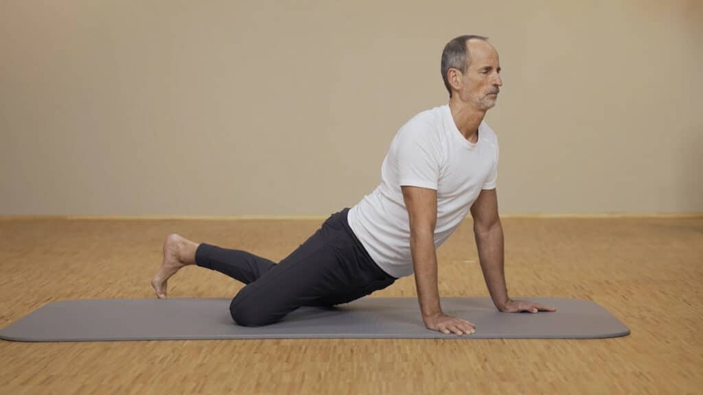 Zu sehen ist Roland Liebscher-Bracht in einem weißen Shirt und schwarzer Trainingshose, der auf einer Übungsmatte eine Übung gegen Schmerzen am unteren Rücken durchführt