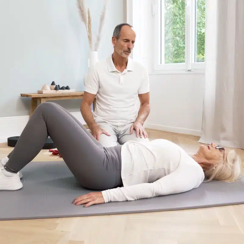 Roland Liebscher-Bracht kniet am Boden neben einer Frau, die sich auf einer Übungsmatte auf den Rücken gelegt und ihre Beine aufgestellt hat.