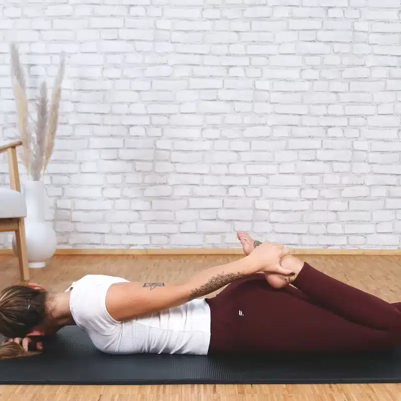 Eine Frau liegt auf dem Bauch. Ihr linkes Bein ist im Knie angewinkelt. Die Frau streckt den linken Arm nach hinten und greift mit ihrer Hand den linken Fuß.