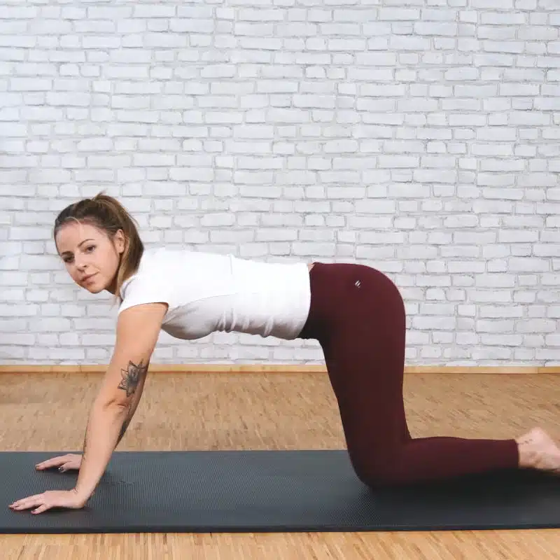 Eine junge Frau ist im Vierfüßlerstand am Boden auf einer schwarzen Yogamatte.