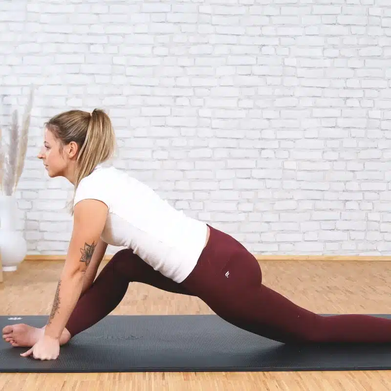Eine junge Frau macht eine Dehnübung auf einer schwarzen Yogamatte. Dazu hat die Frau ihr linkes Bein nach hinten gerade ausgestreckt, während sie das rechte Bein im Knie angewinkelt hat und vor sich hält. Sie stützt ihren Oberkörper mit beiden Armen vom Boden ab.