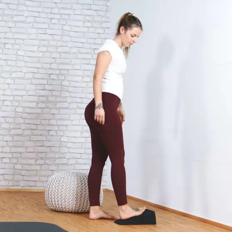 Eine junge Frau steht in Trainingskleidung vor einer weißen Wand. Sie schaut nach unten und platziert ihren rechten Fuß auf dem Knieretter von Liebscher und Bracht.