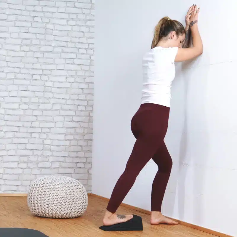 Eine junge Frau dehnt ihre Wade mit dem Knieretter von Liebscher und Bracht. Dazu hat sie ihren rechten Fuß auf dem Knieretter abgestellt und einen Schritt nach vorne gemacht, sodass ihr rechtes Bein nach hinten gerade ausgestreckt ist.