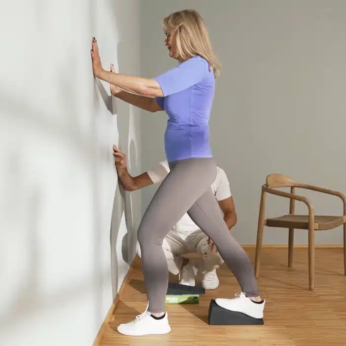 Eine Person steht vor einer Wand und steht mit dem rechten Fuß auf dem Knieretter von Liebscher & Bracht. So wird die Wade gedehnt gegen Knieschmerzen. Das rechte Knie ist vollständig gestreckt und mit den Händen stützt sie sich an der Wand ab.