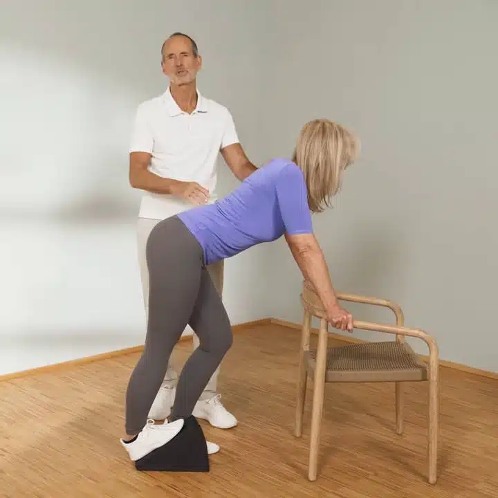 Eine Person steht vor einem Stuhl. Sie hält sich an der Stuhllehne fest und hat den rechten Fuß auf dem Knieretter aufgestellt. Das rechte Bein ist vollständig durchgestreckt und die Person lehnt sich mit dem Oberkörper nach vorne.