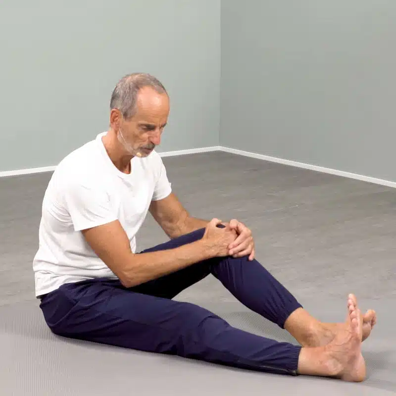 Roland Liebscher-Bracht sitzt am Boden. Sein rechtes Bein liegt ausgestreckt am Boden, während er sich an seinem aufgestellten linken Knie nach vorne zieht.