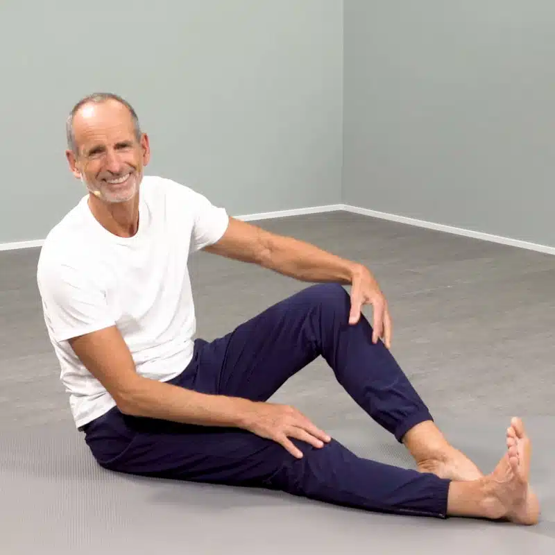 Roland Liebscher-Bracht sitzt am Boden und lächelt in die Kamera. Sein rechtes Bein liegt ausgestreckt vor ihm und sein linkes Bein ist angewinkelt.