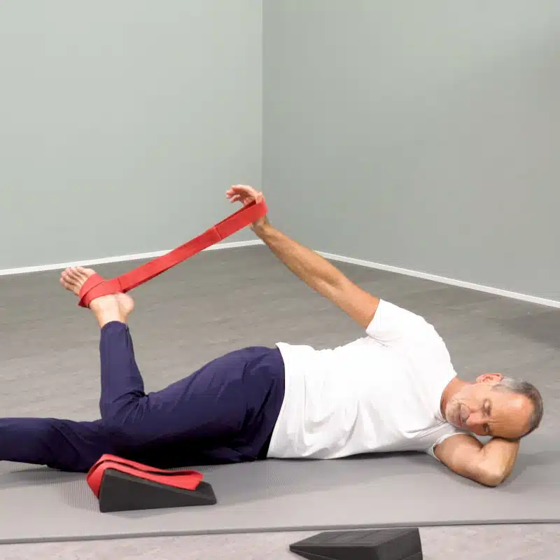 Roland Liebscher-Bracht liegt seitlich auf einer grauen Übungsmatte. Mithilfe einer roten Schlaufe zieht er seinen Fuß zu sich heran. Neben ihm steht ein Knieretter auf Höhe seines Oberschenkels.,