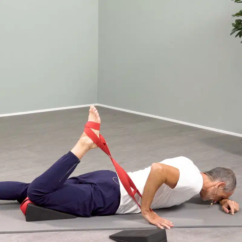 Roland Liebscher-Bracht liegt in Bauchlage am Boden und macht eine Dehnübung. Dazu hat er Oberschenkel und Knie auf dem Knieretter platziert und zieht mithilfe einer roten Schlaufe seinen rechten Fuß zum Gesäß.