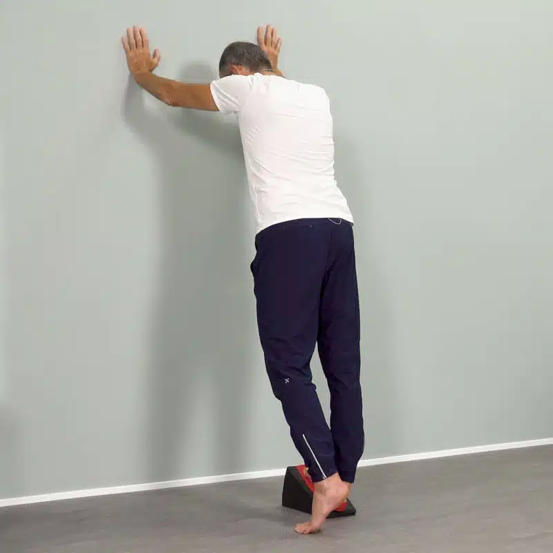 Ein Mann steht mit seinem rechten Fuß auf dem Knieretter von Liebscher & Bracht. Mit dem linken Fuß steht er auf Zehenspitzen und stützt sich nach vorne mit beiden Armen an einer Wand ab.