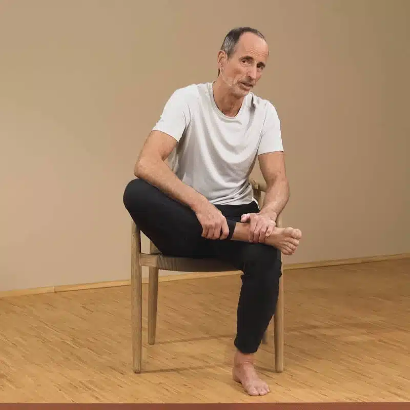 Eine Person sitzt auf einem Stuhl und hat das rechte Sprunggelenk auf dem linken Knie abgelegt. Das rechte Knie zeigt nach rechts und wird mit dem Ellenbogen noch etwas hinunter gedrückt. Beide Hände umgreifen das rechte Sprunggelenk.