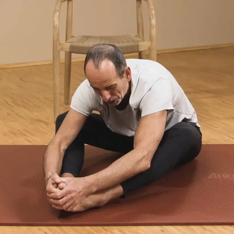 Eine Person sitzt auf einer Matte und hat die Beine vor sich angewinkelt abgelegt. Die Fußsohlen berühren sich und die Knie sind nach rechts und links angewinkelt. Die Person umgreift die Füße mit beiden Händen und lehnt sich nach vorne.