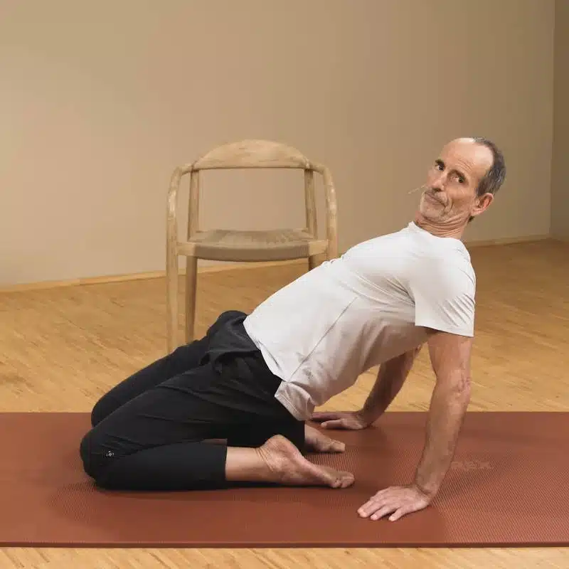 Eine Person sitzt auf dem Rückenretter auf einer Matte. Die Beine sind beide nach hinten angewinkelt. Sie lehnt sich zurück und hat die Hände hinter den Füßen aufgestellt, um sich abzustützen.