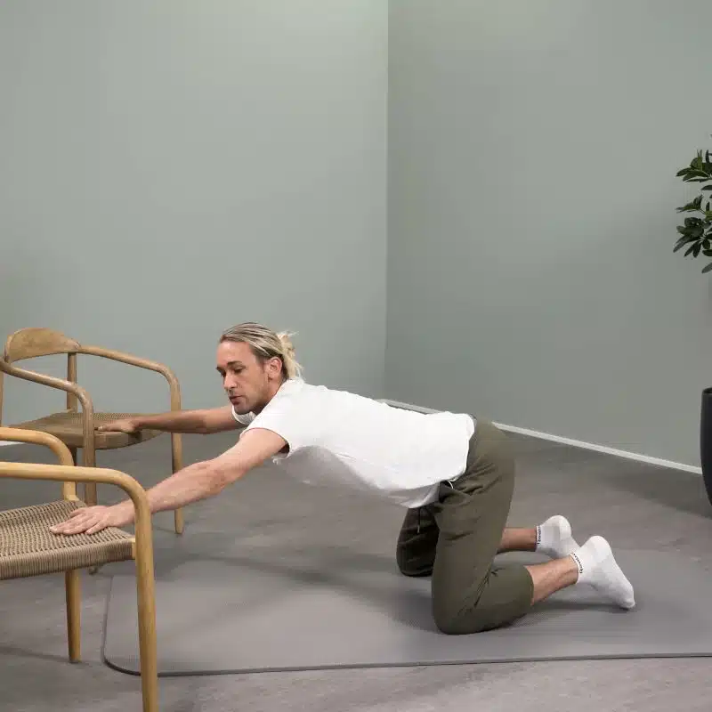 Ein junger Mann kniet vor zwei Stühlen. Er hat seinen Oberkörper vorgebeugt und stützt sich mit ausgestreckten Armen auf den Sitzflächen der beiden Stühle ab.