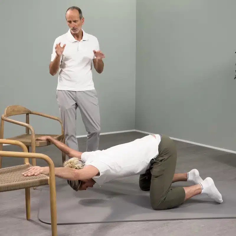 Ein junger Mann kniet vor zwei Stühlen. Er hat seinen Oberkörper weit vorgebeugt, sodass er mit dem Gesicht zum Boden schaut. Mit ausgestreckten Armen hält er sich an den zwei Stühlen fest.