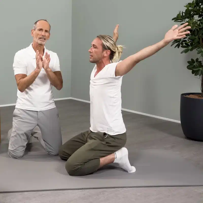 Ein junger Mann kniet am Boden, sein Oberkörper ist gerade und er hat seine Arme seitlich nach oben ausgestreckt.