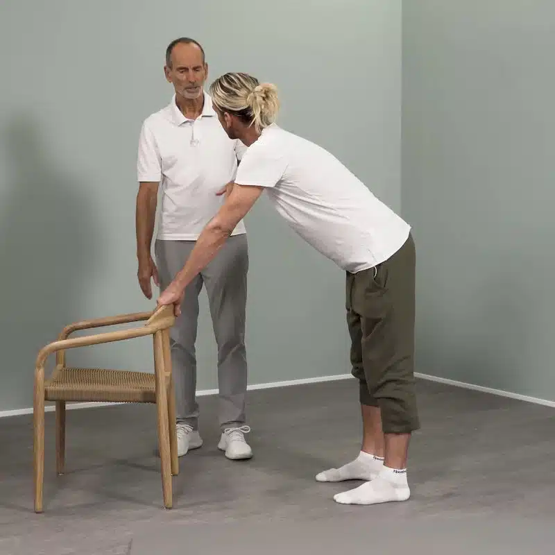 Ein junger Mann steht hinter einem Stuhl und hält sich mit beiden Armen an der Rückenlehne des Stuhlst fest.