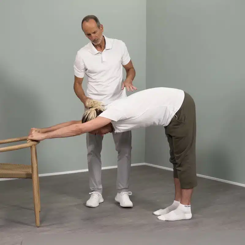 Ein junger Mann steht mit einigem Abstand hinter einem Stuhl. Der Mann hat sich mit seinem Oberkörper weit vorgebeugt und seine Arme über die Schulter nach oben gerade ausgestreckt, sodass er den Stuhl an der Rückenlehne erreicht und sich daran festhalten kann.