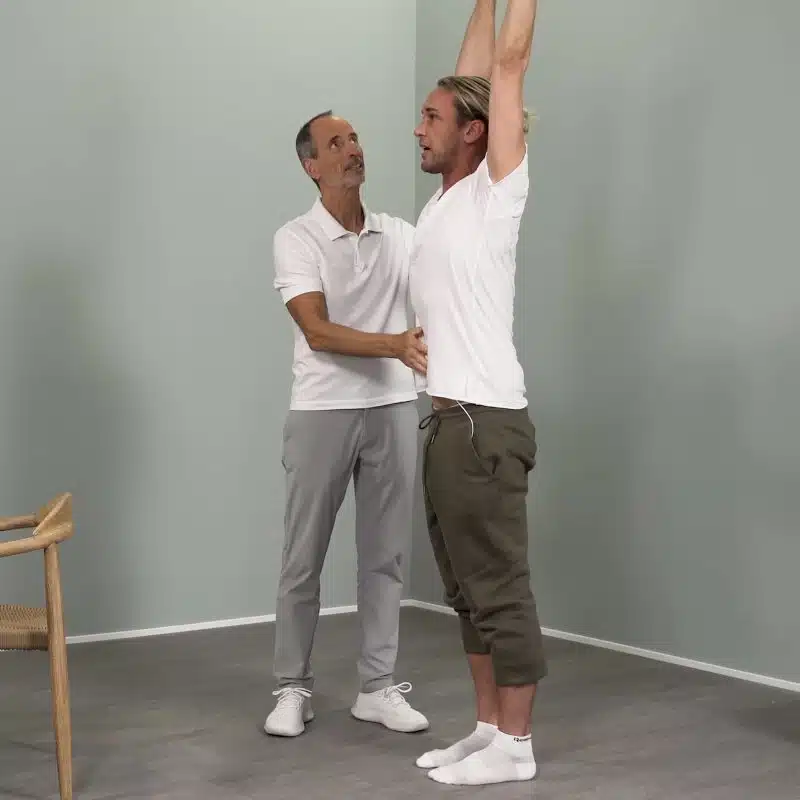 Ein junger Mann steht aufrecht und streckt beide Arme über seinen Kopf nach oben. Neben dem Mann steht Roland Liebscher-Bracht und schaut nach oben.