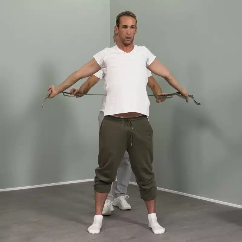 Ein junger Mann hat die Arme seitlich hinter seinem Rücken ausgestreckt und hält mit beiden Händen den Schulterretter von Liebscher & Bracht.