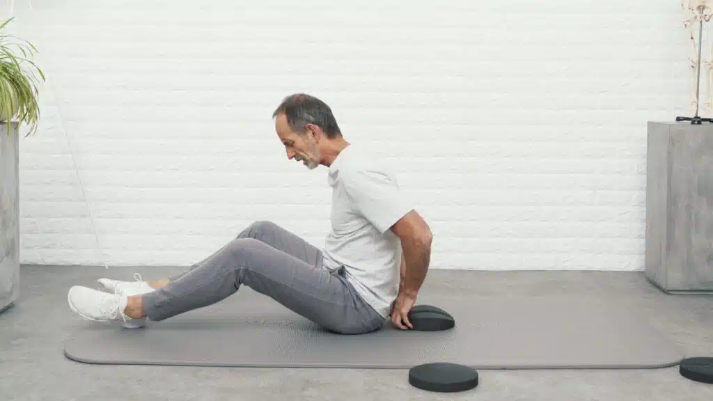 Roland Liebscher-Bracht sitzt auf einer Matte und hat die Beine vor dem Körper aufgestellt. Hinter seinem Rücken platziert er den Rückenretter mit der Einkerbung in der Mitte.