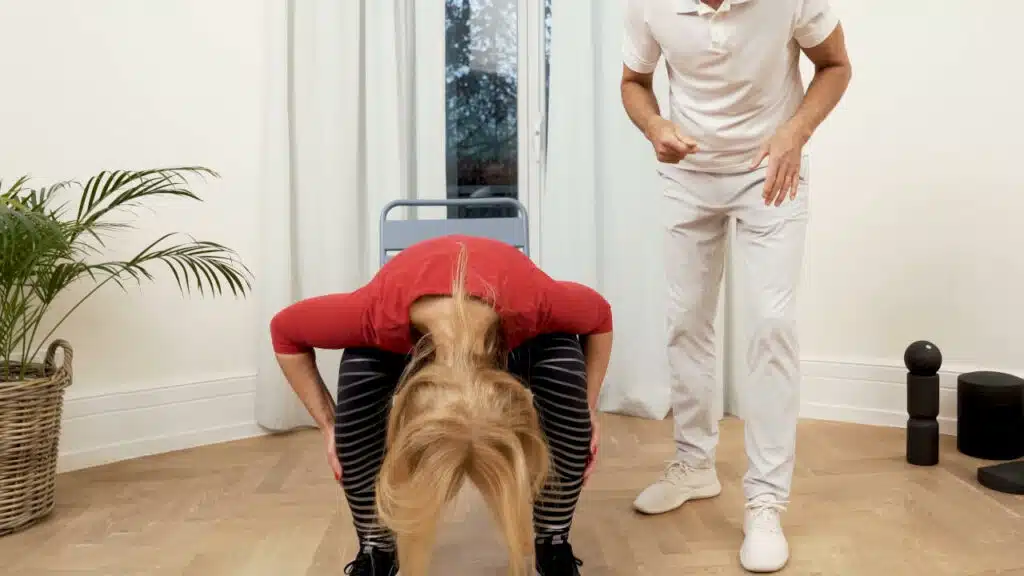 Eine Person sitzt auf einem Stuhl und beugt sich ganz tief nach unten. Ihr Kopf hängt zwischen ihren Knien und die Hände hat sie rechts und links an den Waden abgestellt.