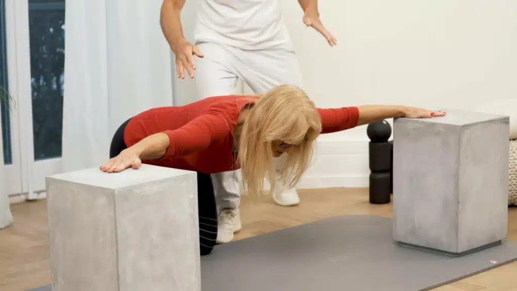 Eine Person kniet auf einer Matte. Die Hände hat sie rechts und links auf zwei Gegenstände, etwa auf Schulterhöhe abgelegt. Ihre Arme bilden dadurch eine lange Line. Sie lässt den Brustkorb nach unten hängen. Der Kopf ist in gerader Linie mit der Wirbelsäule