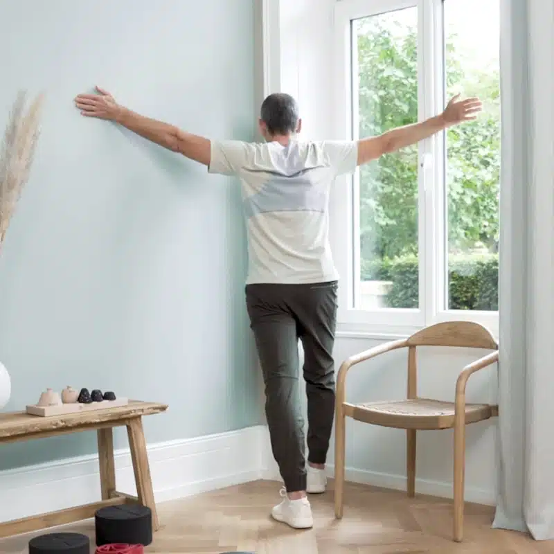 Roland Liebscher-Bracht steht mit ausgebreiteten Armen in einer Raumecke und dehnt den Brustkorb