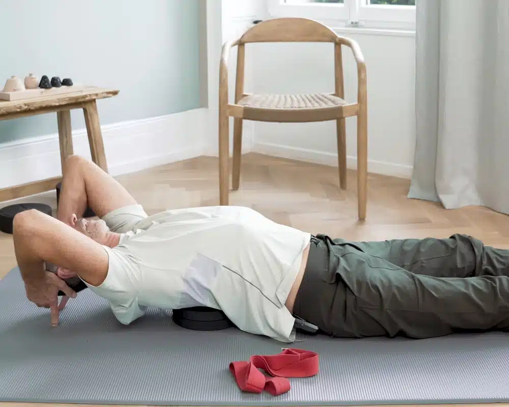 Roland Liebscher-Bracht macht eine Übung mit dem Rückenretter, indem er diesen auf dem Boden liegend unter den Rücken legt und die Arme Richtung Kopf zieht.