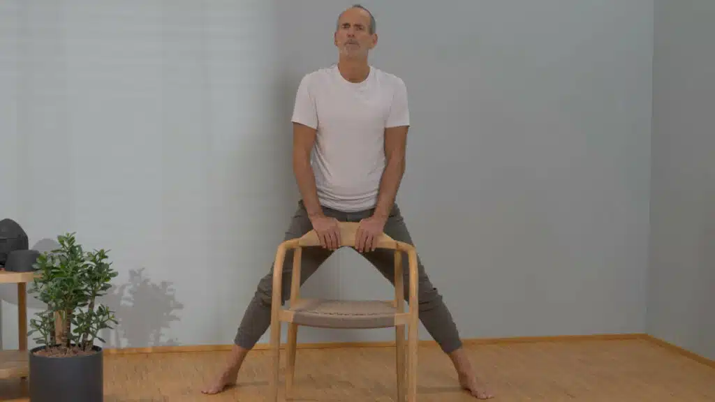 Roland Liebscher-Bracht steht vor einem Stuhl und hält sich mit beiden Händen an der Lehne des Stuhls fest. Sein Oberkörper ist aufrecht, die Beine hat er weit geöffnet.