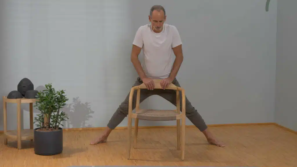 Roland Liebscher-Bracht steht vor einem Stuhl und hält sich mit beiden Händen an der Lehne des Stuhls fest. Sein Oberkörper ist aufrecht, die Beine hat er weit geöffnet. Seine Füße zeigen leicht nach außen.