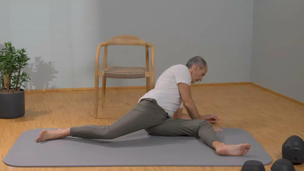 Roland Liebscher-Bracht sitzt auf dem Boden und hat das linke Bein vor sich angewinkelt. Das rechte Bein liegt lang nach hinten gestreckt. Den Oberkörper neigt er nach links und stützt sich mit den Händen neben dem linken Knie ab.
