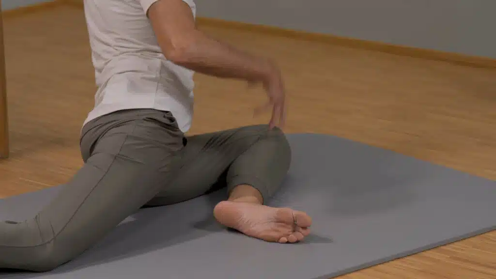 Der Ausschnitt einer Gesäßdehnung zeigt das Becken und die Beine von Roland Liebscher-Bracht. Das linke Knie ist vor ihm angewinkelt, er deutet mit der Hand darauf. Das rechte Bein ist nach hinten gestreckt und bis zum Knie abgebildet.