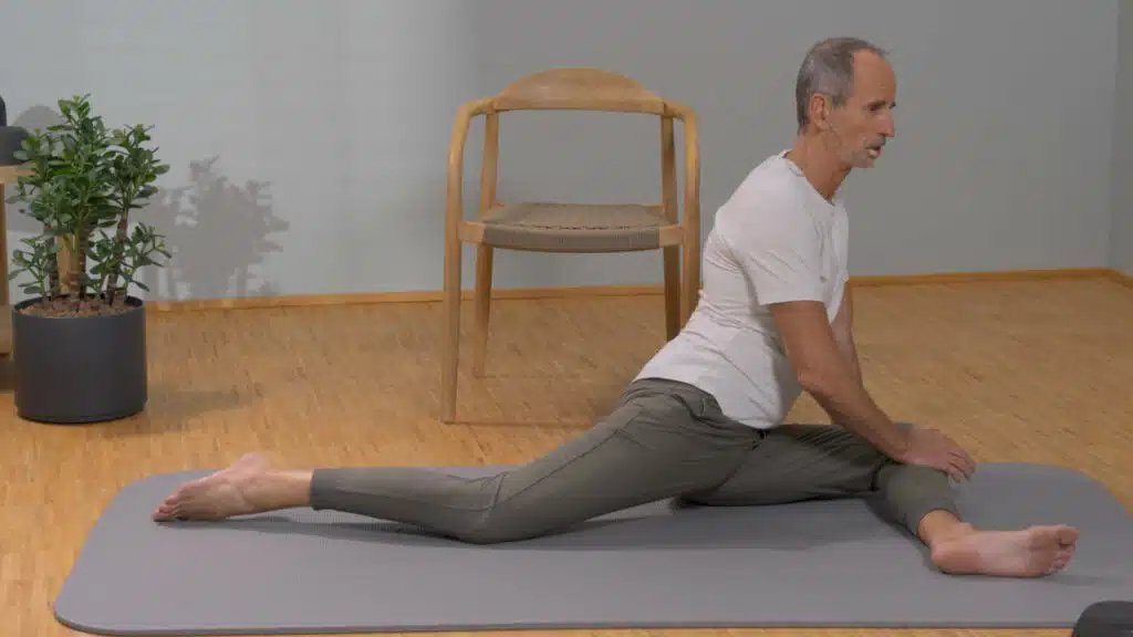 Roland Liebscher-Bracht sitzt auf dem Boden und hat das linke Bein vor sich angewinkelt und das rechte Bein nach hinten ausgestreckt. Der Oberkörper ist aufrecht.