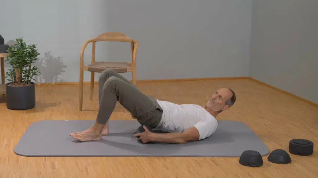 Roland Liebscher-Bracht schiebt sich den ISG-Ischias-Retter unter das linken Gesäß. Die Füße und die Ellenbogen sind am Boden aufgestellt.