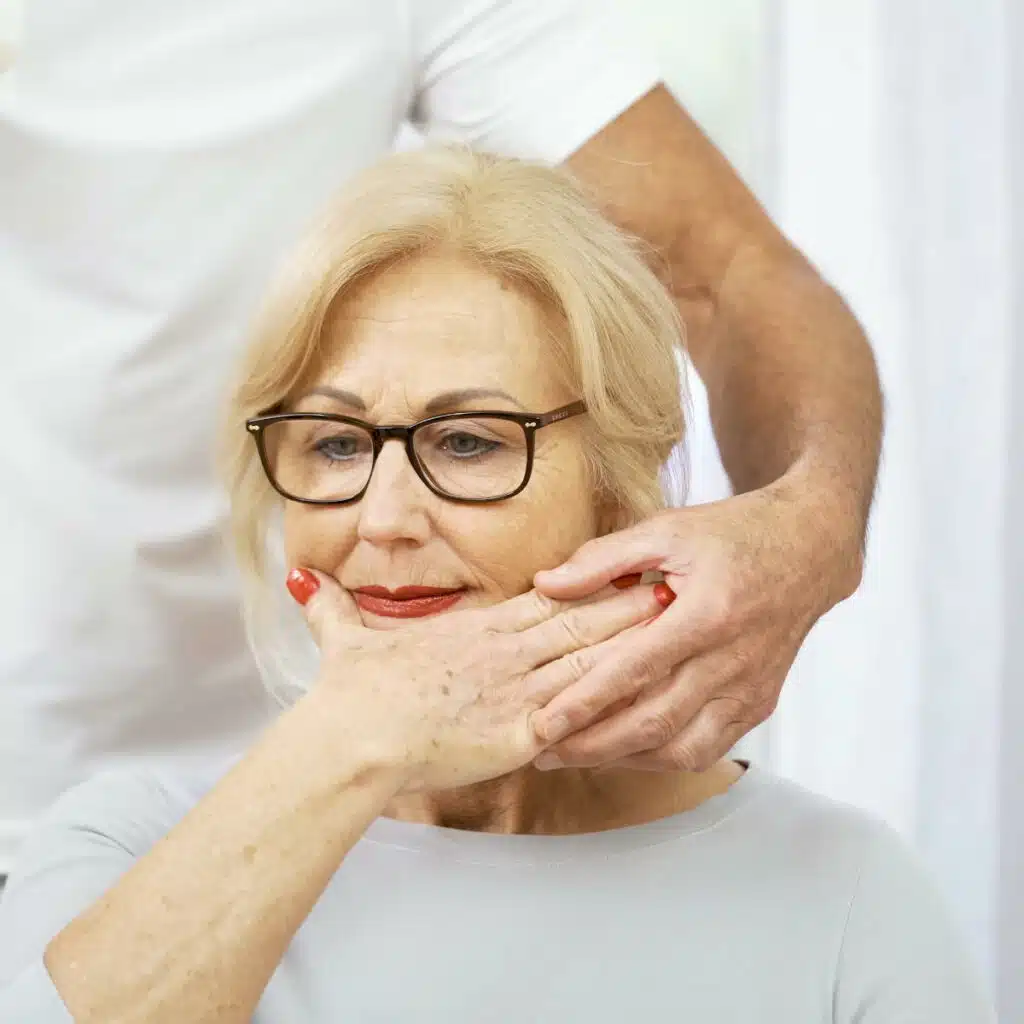 Eine Person hat die rechte Hand zum Gesicht gezogen und den Daumen von den anderen Fingern abgespreizt. Sie legt die Hand auf ihr Kinn, direkt unterhalb des Mundes. Roland Liebscher-Bracht hilft ihr dabei und führt ihre Hand.