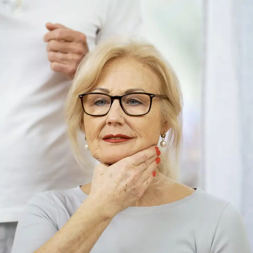 Eine Person hat die rechte Hand zum Gesicht gezogen und den Daumen von den anderen Fingern abgespreizt. Sie legt die Hand um den Unterkiefer, direkt unterhalb des Mundes.