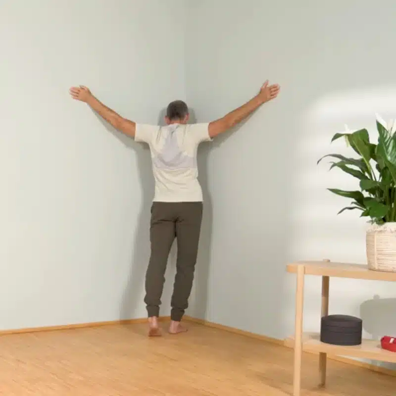 Roland Liebscher-Bracht steht mit seitlich nach oben gestreckten Armen in einer Raumecke.