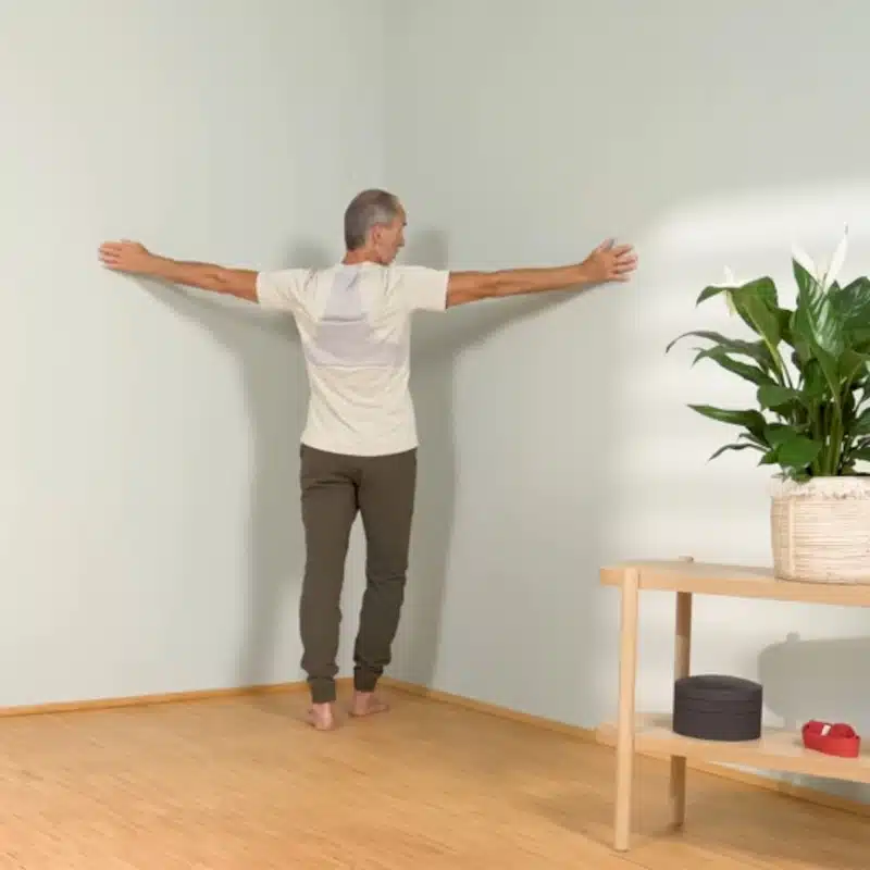 Roland Liebscher-Bracht steht mit seitlich nach oben gestreckten Armen in einer Raumecke und richtet sich für eine Dehnung aus