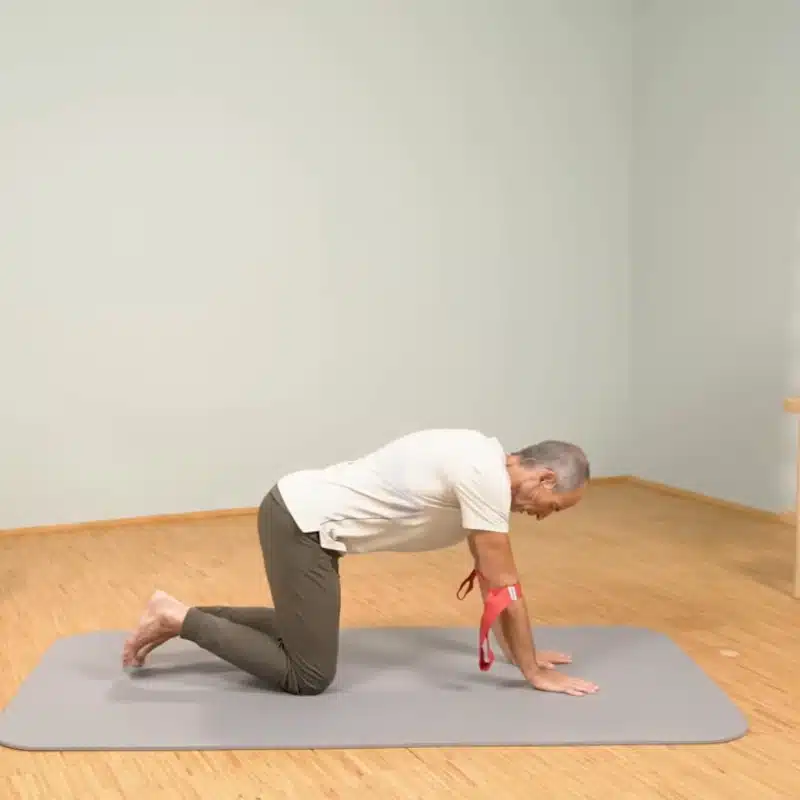 Roland Liebscher-Bracht kniet im Vierfüßlerstand auf dem Boden und hat eine rote Übungsschlaufe um seine Arme gebunden