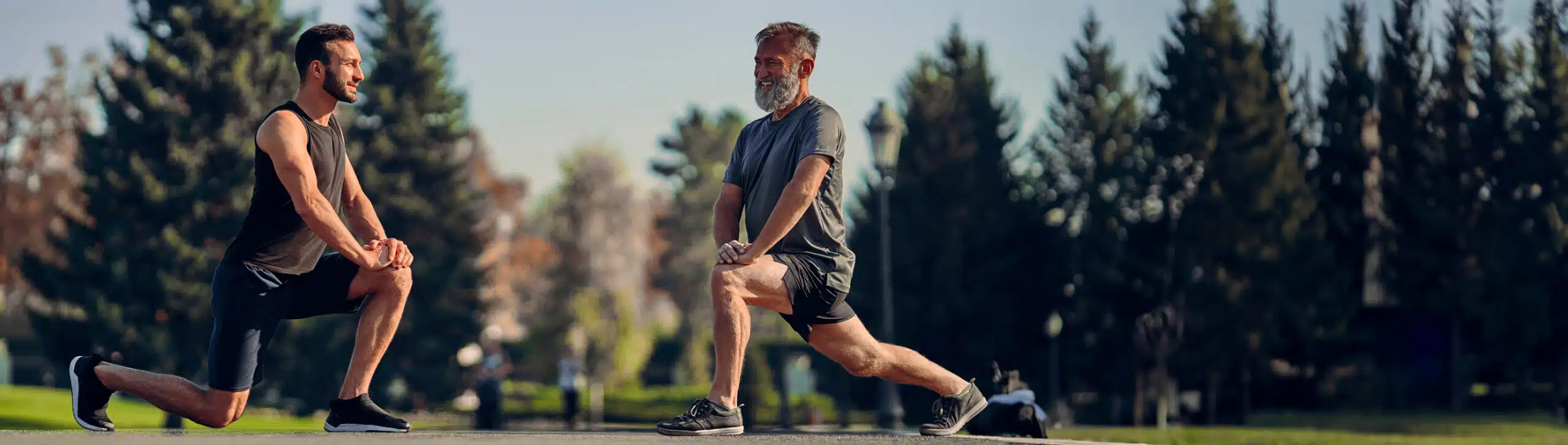 alter Mann und junger Mann machen eine Dehnübung der Beine: sie stehen in einem weiten Ausfallschritt, vorderes Knie im 90Grad-Winkel angebeugt, das hintere Bein ausgestreckt