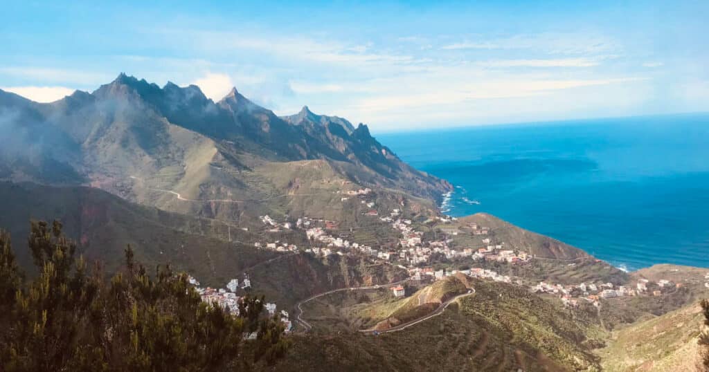 Fernblick auf ein Tal mit Häusern, dahinter hoch aufragende Berge und das Meer