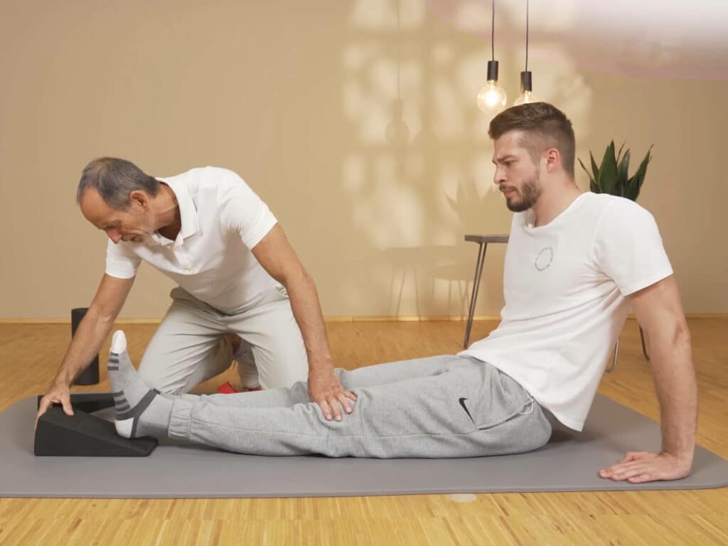 Ein Mann sitzt mit ausgestreckten Beinen am Boden und legt einen Fuß auf dem Knieretter von Liebscher & Bracht ab.