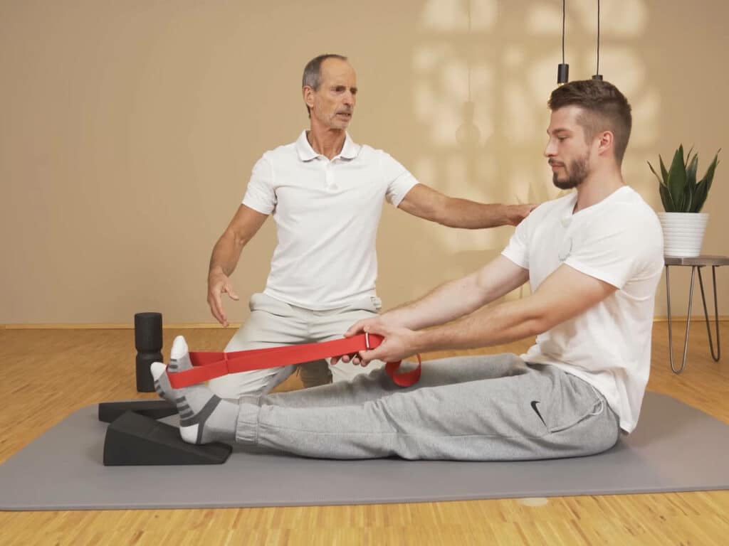Ein Mann sitzt am Boden. Er hat sein ausgestrecktes Bein auf dem Knieretter abgelegt und zieht mithilfe einer roten Übungs-Schlaufe den hochgelegten Fuß zu sich heran.