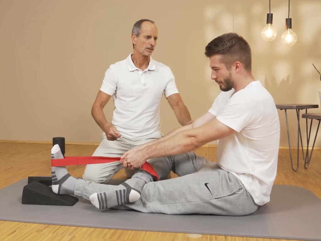 Ein Mann sitzt am Boden und macht eine Übung gegen Knieschmerzen. Sein linkes Bein ist ausgestreckt und liegt leicht erhöht auf einem Knieretter. Sein rechtes Bein ist angewinkelt und liegt auf dem linken Knie. Der Mann sitzt gerade und zieht sich mit beiden Armen an einer roten Schlaufe nach vorne in Richtung seines linken Fußes.