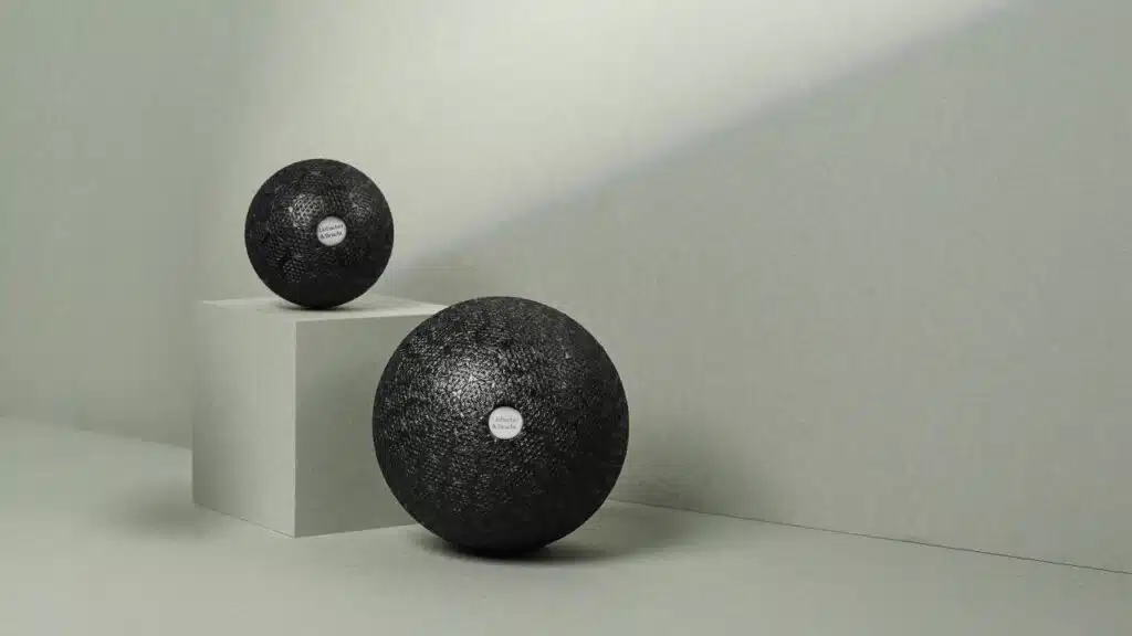 Zwei schwarze Faszienbälle von Liebscher & Bracht liegen vor einem hellgrauen Hintergrund. Rechts liegt ein größerer Faszienball und links liegt eine kleinere Faszienkugel leicht erhöht auf einer grauen Box.