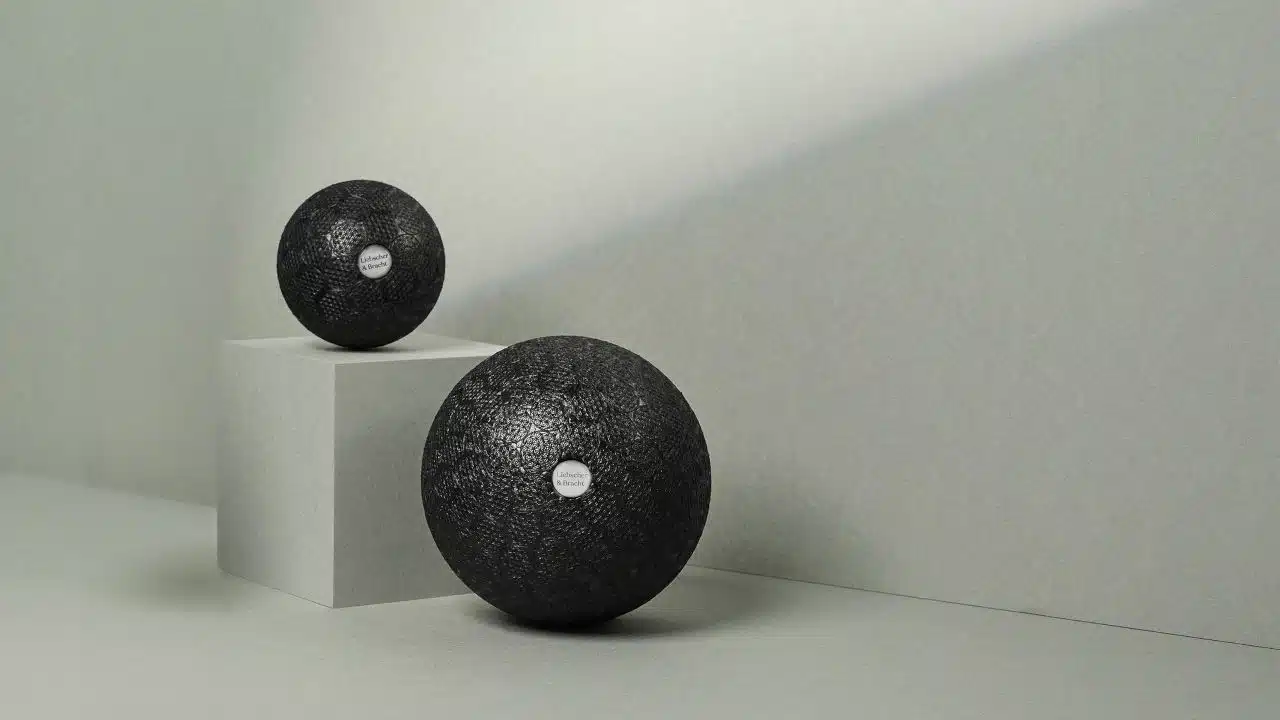 Zwei schwarze Faszienbälle von Liebscher & Bracht liegen vor einem hellgrauen Hintergrund. Rechts liegt ein größerer Faszienball und links liegt eine kleinere Faszienkugel leicht erhöht auf einer grauen Box.