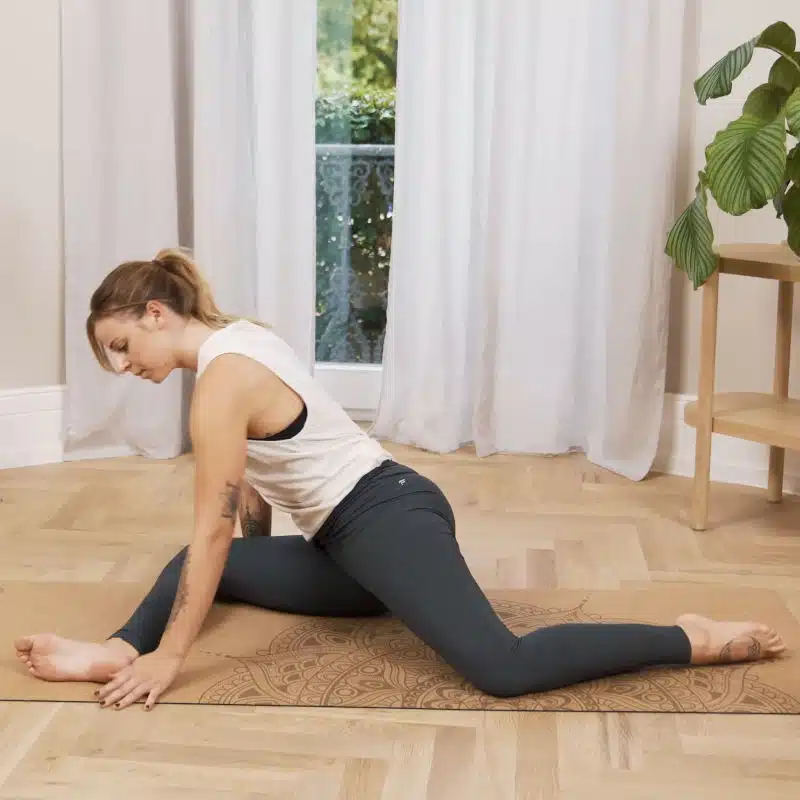 Eine Frau bereitet sich am Boden auf eine Übung vor. Sie hat dazu ihr rechtes Bein angewinkelt vor sich abgelegt und stützt sich mit beiden Händen am Boden ab.