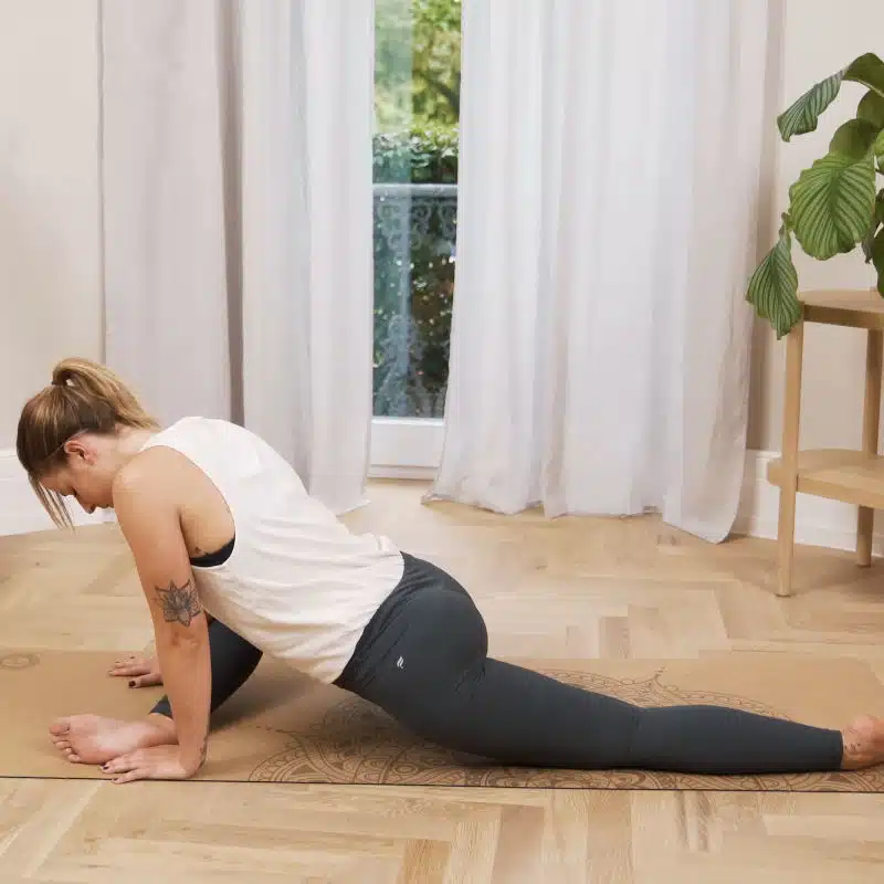 Eine Frau macht am Boden eine Übung. Dazu hat sie eine Position eingenommen, in der ihr linkes Bein vollständig nach hinten ausgestreckt ist, während ihr rechtes Bein angewinkelt vor ihr liegt. Außerdem beugt sie sich mit dem Oberkörper über das vordere Bein und stützt sich mit beiden Händen am Boden ab.