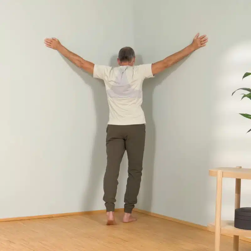 Roland Liebscher-Bracht zeigt eine Dehnübung in einer Raumecke. Seine Arme sind seitlich nach oben ausgestreckt.